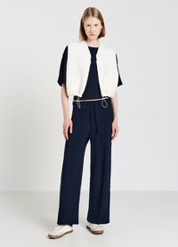 Weiß gesteppte Weste über einem navyfarbenen T-Shirt, kombiniert mit navyblauen, weit geschnittenen Hosen. Das Model trägt weiße Sneakers mit brauner Sohle und hat einen neutralen Hintergrund.