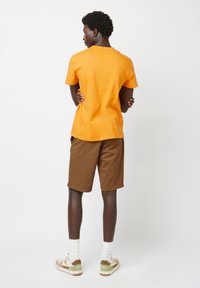 T-shirt in cotone arancione, maniche corte, scollatura rotonda, abbinato a pantaloni corti marroni. Dettagli con spille da balia sul retro. Indossato con calzini bianchi e sneaker.