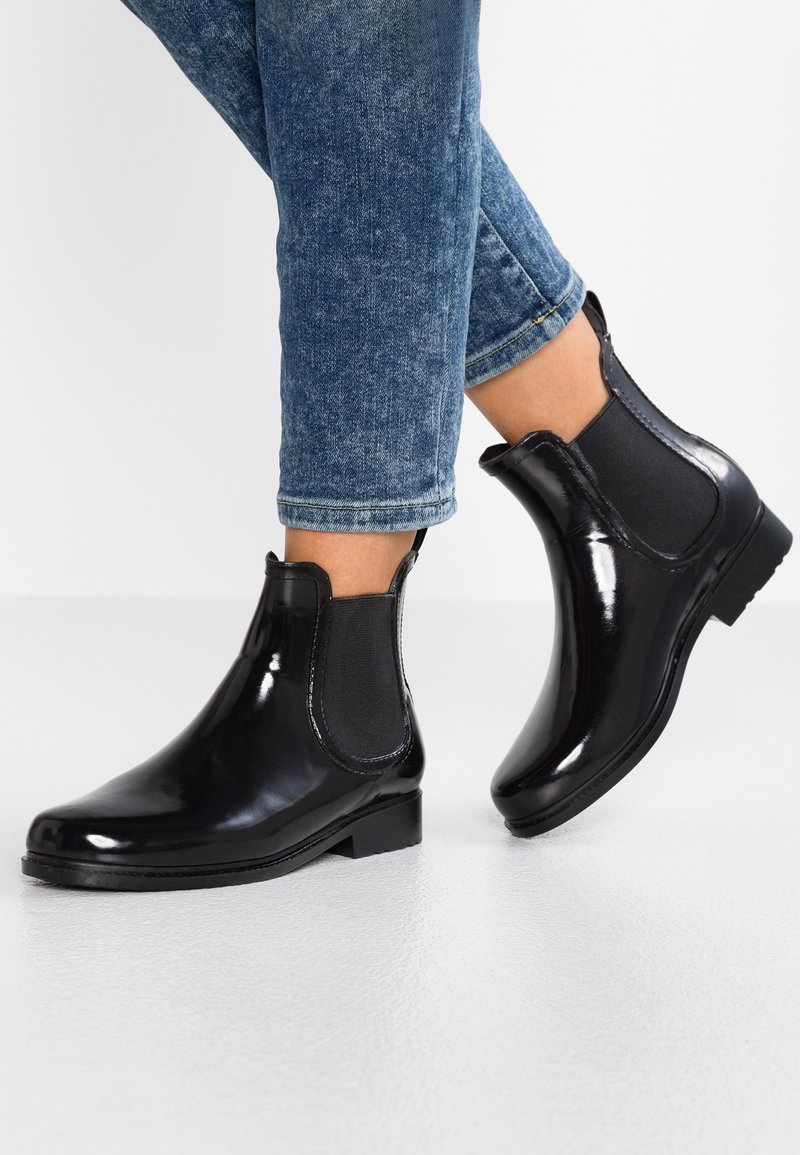 Personne portant des bottines Chelsea noires brillantes avec des panneaux latéraux élastiques et un jean bleu retroussé sur une surface blanche.