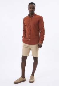Camisa de botones de color óxido con mangas largas, combinada con pantalones cortos color beige y zapatillas beige. Textura suave, detalle sutil del logo en la camisa.