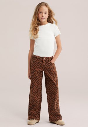 Jeune fille aux longs cheveux ondulés portant un haut blanc à manches courtes, un pantalon large marron à rayures tigre et des chaussures beiges, debout avec une main dans la poche.