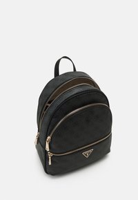 Mochila mini negra de Guess con tres compartimentos con cremallera, patrón de logo sutil y cremalleras y placa de logo en tono dorado sobre fondo blanco.