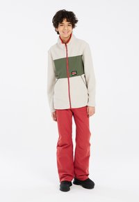 Chaqueta de forro polar en blanco roto con panel verde oliva, cremallera roja y parche de logo; combinada con pantalones rojos de corte holgado y botas negras.