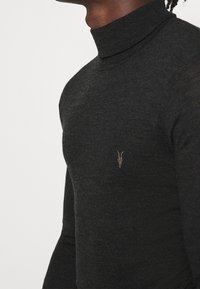 Schwarzer Rollkragenpullover aus glattem Strickmaterial; taillierte Passform mit einem kleinen braunen Ziegensymbol auf der linken Brust.