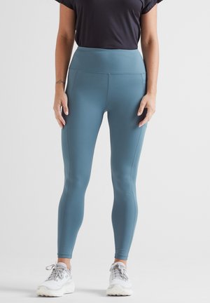 Blue high-waisted leggings with a smooth texture and seam detailing, paired with a black short-sleeve top and white sneakers.