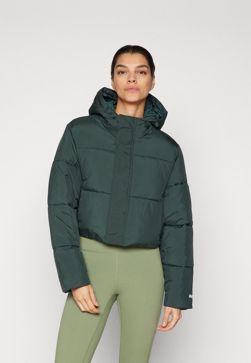Veste matelassée courte vert foncé avec une capuche, présentant une fermeture éclair sur le devant, une texture matelassée et des poignets côtelés. Associée à des leggings verts olive.