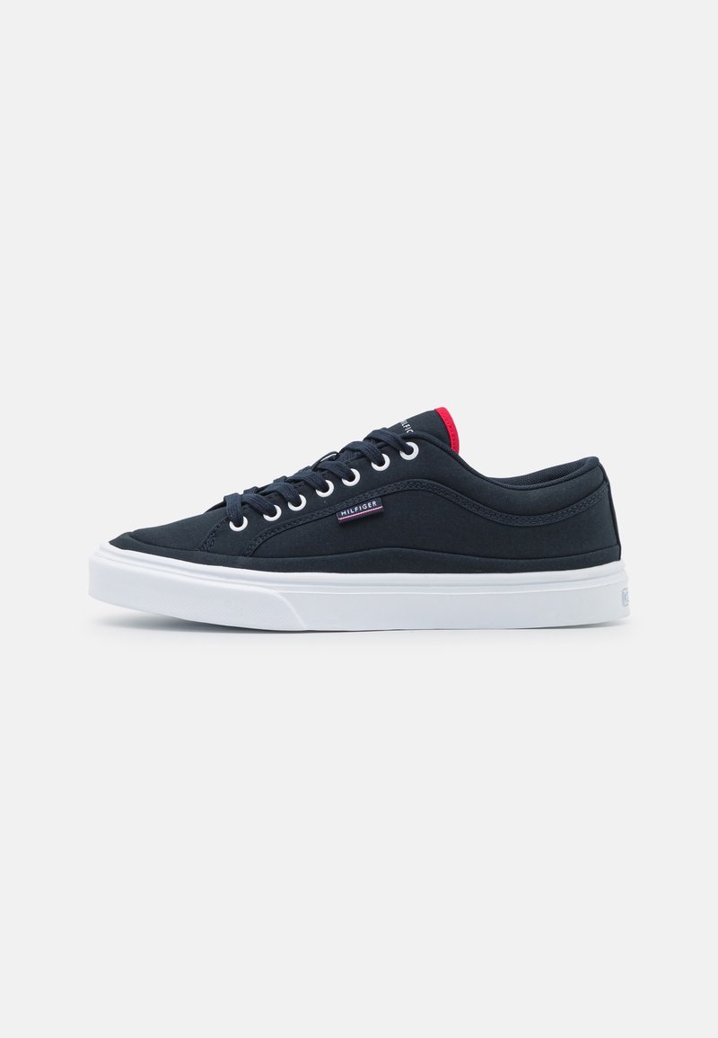 Baskets basses noires avec semelle blanche, lacets noirs, petite étiquette en tissu avec logo et accent rouge sur la languette, présentées sur fond blanc.