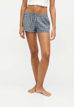 Kvinde iført sort-hvide ternede shorts og en hvid cropped top, stående barfodet mod en ensfarvet baggrund.