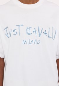 Baltos medvilnės marškiniai su apskrito kaklo iškirpte. Puošia mėlyna įsiuvinėta užrašas: "Just Cavalli Milano" pateiktas laisvalaikio stiliumi.