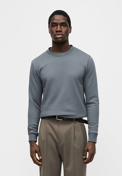 Άνδρας με σκουρόχρωμο δέρμα που φοράει γκρι μακρυμάνικο πουκάμισο και μπεζ πτυχωτό παντελόνι με καφέ πλεκτή ζώνη, στέκεται μπροστά σε απλό φόντο.