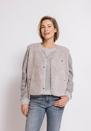 Woman with short blond hair wearing a gray fleece vest over a gray sweatshirt and blue jeans, standing with one hand on her hip.