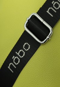 Grünes strukturiertes Material mit einem schwarzen Riemen, der mit silbernen Beschlägen und dem "nabo"-Logo versehen ist. Das Design hebt die glatte Oberfläche und die kontrastierenden Farben hervor.