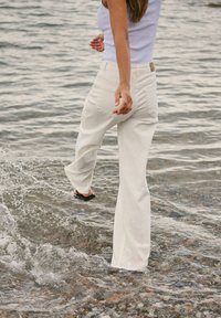 Personne en pantalon blanc et débardeur clair marchant dans l'eau peu profonde, éclaboussant, avec des vagues autour des pieds.
