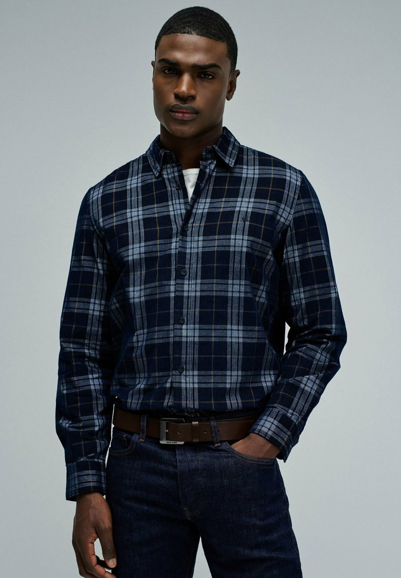 Camisa de cuadros en tonos azul marino y gris, con cuello, mangas largas y botones en la parte delantera. Lleva un cinturón marrón y jeans oscuros.