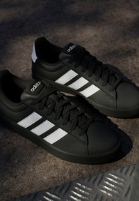 Baskets Adidas noires avec des bandes blanches, des lacets noirs et une semelle en caoutchouc reposant sur une surface texturée dans une lumière douce.