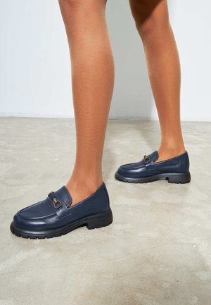 Benen met doorschijnende bruine panty's en marineblauwe leren loafers met zwarte dikke zolen, staand op een betonnen vloer.