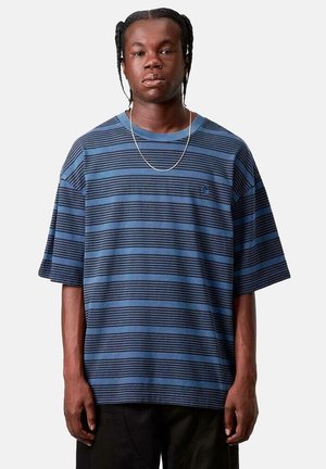 Hombre con cabello trenzado que lleva una camiseta holgada de rayas horizontales azules y negras y pantalones negros, de pie contra un fondo liso.