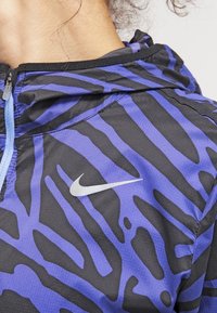 Sudadera de Nike con un patrón de rayas de cebra en negro y morado, con cierre frontal con cremallera y acento del logo plateado en el pecho. Tejido ligero.