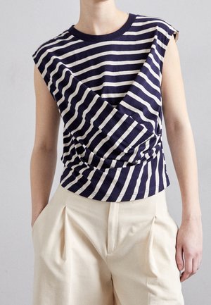 Top sans manches rayé bleu marine et blanc avec un détail de pli diagonal, associé à un pantalon plissé beige taille haute, mains dans les poches.