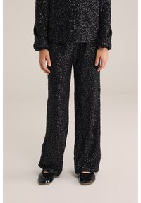 Mustat paljetti-asusteet, joissa on pitkähihainen paita ja löysät housut. Kangas on tekstuurinen ja kiiltävä pinta. Yhdistetty mustiin kenkiin.