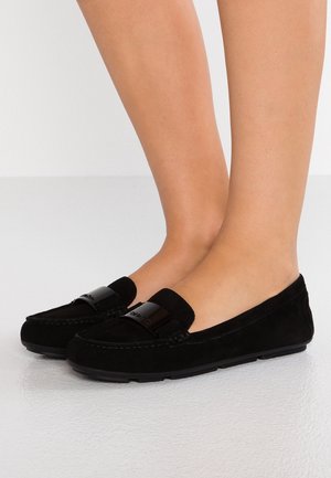 Schwarze Wildleder-Loafers mit niedrigem Absatz und einem glänzenden schwarzen Riemen über dem Spann, getragen an nackten Füßen auf einem hellen Boden.