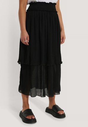 Mujer con falda midi negra en capas con cintura fruncida y sandalias negras de plataforma sin cierre sobre un fondo liso.