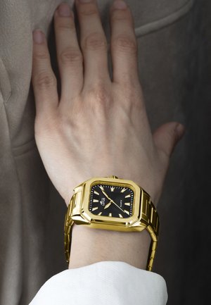 Orologio in metallo dorato con quadrante quadrato, fondale nero e indicatori dell'ora luminosi. Attachato a un bracciale a maglie dorate; polso parzialmente visibile.