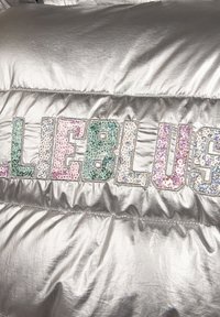 Srebrna puchowa kurtka z opalizującymi cekinowymi literami "ELLIEBLU" w kolorach różowym, niebieskim i zielonym. Gładka, błyszcząca tkanina z pikowanym wzorem.