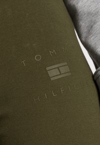 Tkanina w kolorze oliwkowym z subtelnym tłoczonym logo "TOMMY HILFIGER" oraz graficznym emblematem. Tło jest szare.