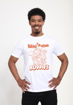 Mann lächelt mit Händen in den Hüften, trägt ein weißes T-Shirt mit orangefarbenem Schriftzug „Bikini Bottom Buddies“ und Comicfiguren aus SpongeBob Schwammkopf.