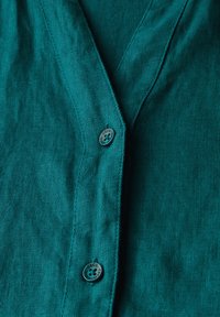 Teal linnekjort med knapp framtill och V-ringning, med ton-i-ton sömmar och en rund knapp märkta "CECIL." Texturen ser mjuk och lätt ut.