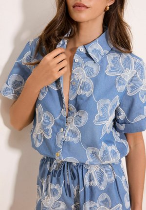 Nő, kék rövid ujjú gombos ingben és hozzáillő, fehér nagy virágmintás megkötős rövidnadrágban, kezében tartja az ing gallérját.