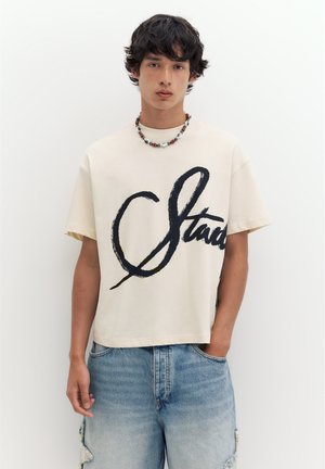 Młody mężczyzna o ciemnych włosach, ubrany w beżowy t-shirt z czarnym napisem i niebieskie dżinsy, stojący na białym tle.