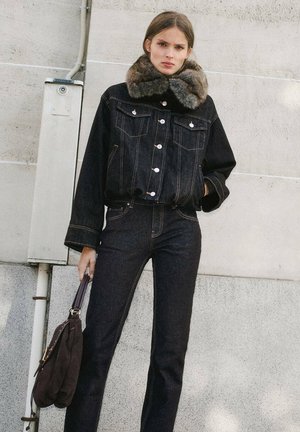 Une femme porte une veste en jean foncé à grand col en fourrure, assortie à un jean, et tient un sac à main marron devant un mur en béton.