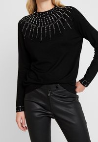 Schwarzer Pullover mit silbernen Strass-Verzierungen, die in einem Sonnenstrahlenmuster auf den Schultern und Ärmeln angeordnet sind. Stilisiert mit eng anliegenden schwarzen Lederhosen.