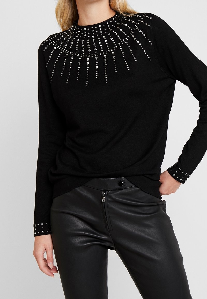 Schwarzer Pullover mit silbernen Strass-Verzierungen, die in einem Sonnenstrahlenmuster auf den Schultern und Ärmeln angeordnet sind. Stilisiert mit eng anliegenden schwarzen Lederhosen.