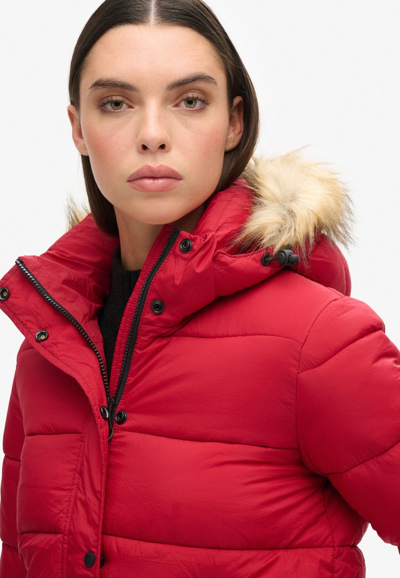 Donna con capelli scuri lisci che indossa un piumino rosso con cappuccio bordato di pelliccia e un maglione nero su sfondo bianco semplice.