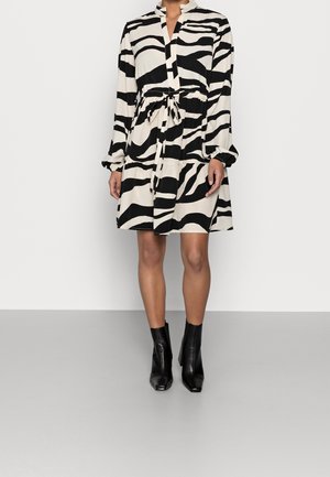 Kobieta w sukience z długim rękawem o czarno-białym wzorze zebry, z taliowaniem i falbanami przy kołnierzu, w połączeniu z czarnymi botkami za kostkę.