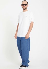 Witte T-shirt met korte mouwen en een klein zwart logo op de borst, gecombineerd met losse blauwe spijkerbroeken en zwarte sneakers.