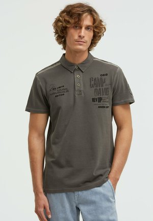 Junger Mann mit lockigem hellbraunem Haar, der ein dunkelgraues Poloshirt mit aufgedrucktem Text und hellblaue Jeans trägt, steht vor einfarbigem Hintergrund.