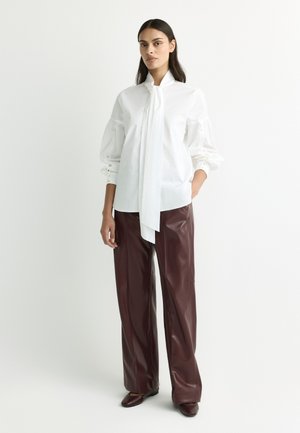 Woman wearing a white blouse with a long front tie and brown leather wide-leg pants standing against a plain light background.