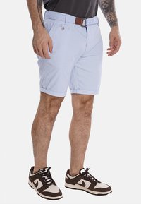 Pantaloni corti di tessuto intrecciato blu chiaro con risvolti, passanti per cintura e un dettaglio in pelle marrone. Abbinati a sneaker marroni e bianche.