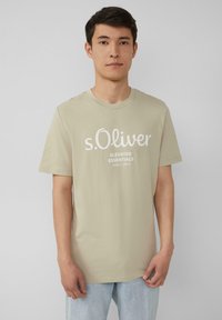 Beige katoenen T-shirt met korte mouwen, ronde halslijn en witte grafische tekst. Draag het met lichtblauwe jeans. Effen achtergrond.