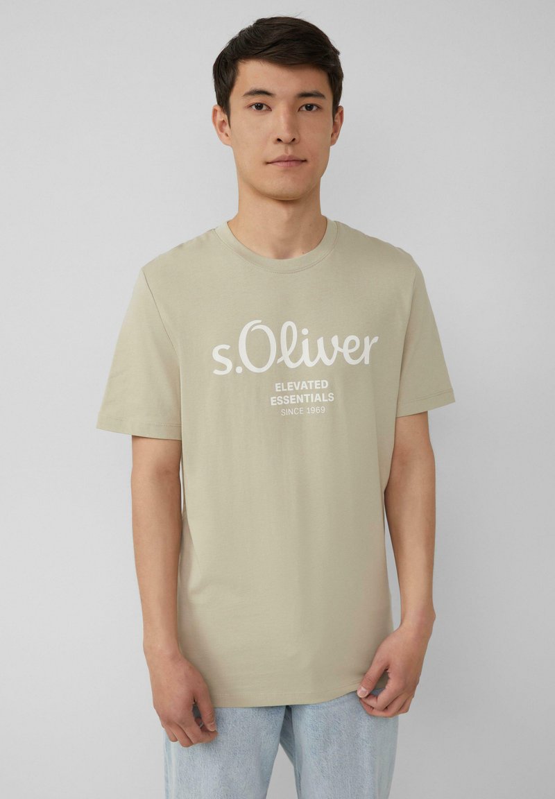Beige katoenen T-shirt met korte mouwen, ronde halslijn en witte grafische tekst. Draag het met lichtblauwe jeans. Effen achtergrond.