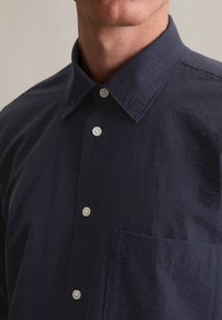 Donkerblauw overhemd met knopen, voorzien van een standaard kraag, een enkele borstzak en witte knopen, met een gladde, licht gestructureerde stof.