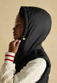 Czarna kamizelka z kapturem i błyszczącymi akcentami, noszona na kremowym swetrze z dzianiny z czerwonymi paskami na mankietach. Gładka faktura i dopasowany krój.