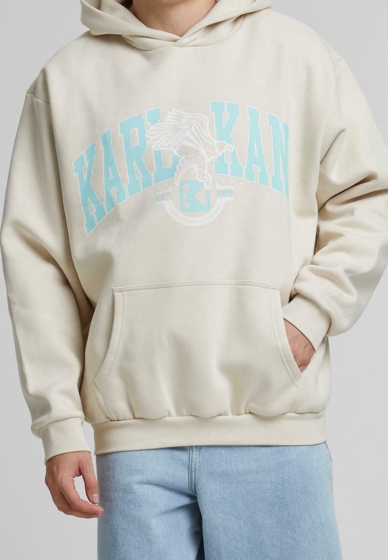 Beige hoodie med en stor, ljusblå tryckt logotyp och örn-design. Den har en framficka och ribbade muddarna vid fållen och ärmarna.