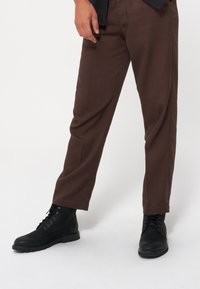 Brązowe dopasowane spodnie o gładkiej fakturze, slim fit, z przeszyciami, zestawione z czarnymi skórzanymi butami na sznurowadła.