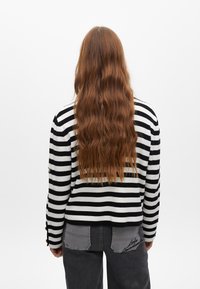 Sweter w pasy w czerni i bieli, z okrągłym dekoltem i długimi rękawami, zestawiony z szarymi spodniami z haftem w kieszeniach.