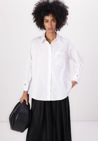 White button-up shirt made of cotton with long sleeves, a chest pocket, and gold-button accents, paired with a black skirt and handbag.